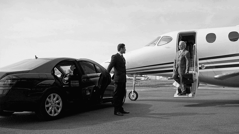 Black and white: traveler leaving a private jet on the tarmac toward a chauffeur and open door of a luxury sedan