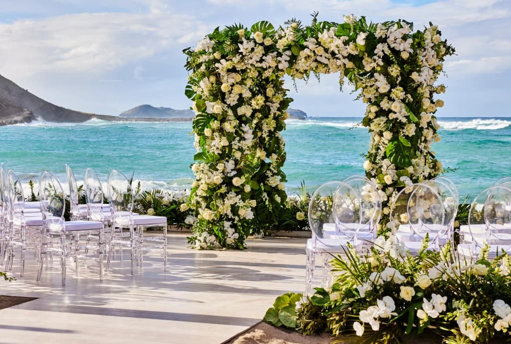 Luxury oceanfront wedding setup: white floral arch, ghost chairs on a deck above turquoise waves and green coastline
