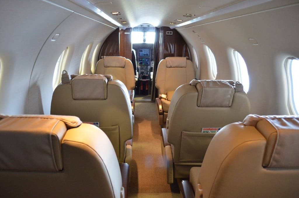 Private jet cabin with tan leather seats, central aisle and view toward the cockpit through bright oval windows